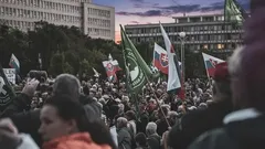 Demonštrácia davu ľudí s vlajkami pred budovami v meste za súmraku.